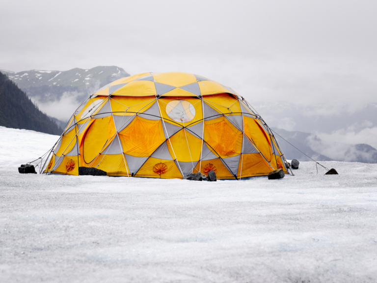 Geodätische Kuppelzelt in Gelb auf einem Gletscher mit nebelverhangenen Bergen im Hintergrund.