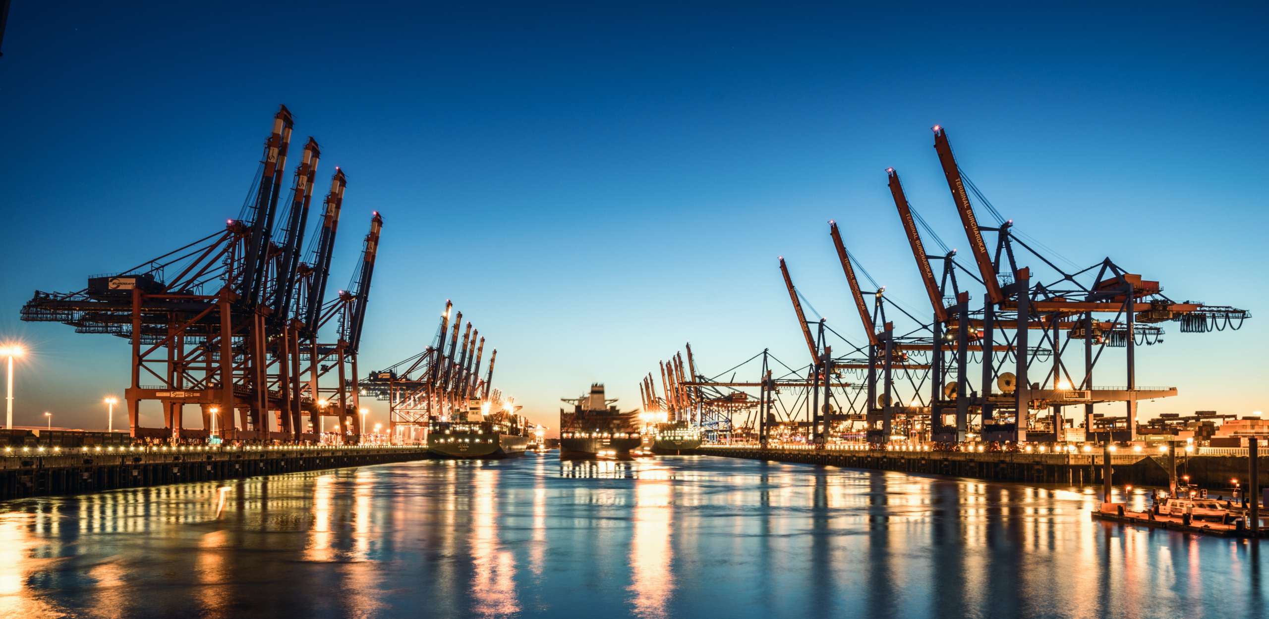 Industriehafen bei Nacht mit erleuchteten Containerkränen und Spiegelung im Wasser.
