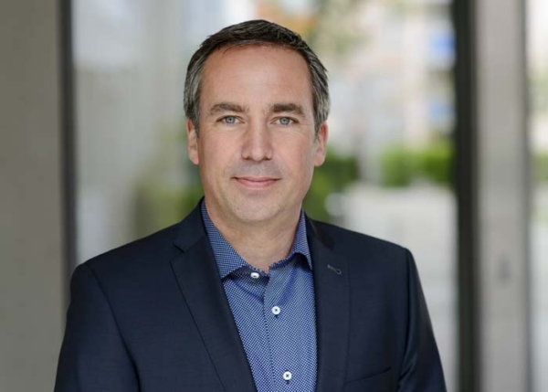 Geschäftsmann mit Anzug und gepunktetem Hemd lächelt vor unscharfem Bürohintergrund.