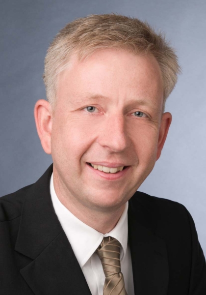 Porträt eines lächelnden Mannes mit kurzen grauen Haaren, Business-Anzug und gestreifter Krawatte.