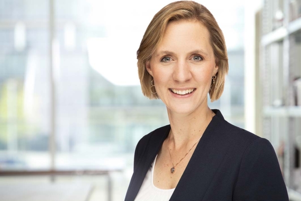 Porträt einer lächelnden Geschäftsfrau mit kurzen blonden Haaren in dunklem Blazer und weißem Top vor unscharfem Bürohintergrund.