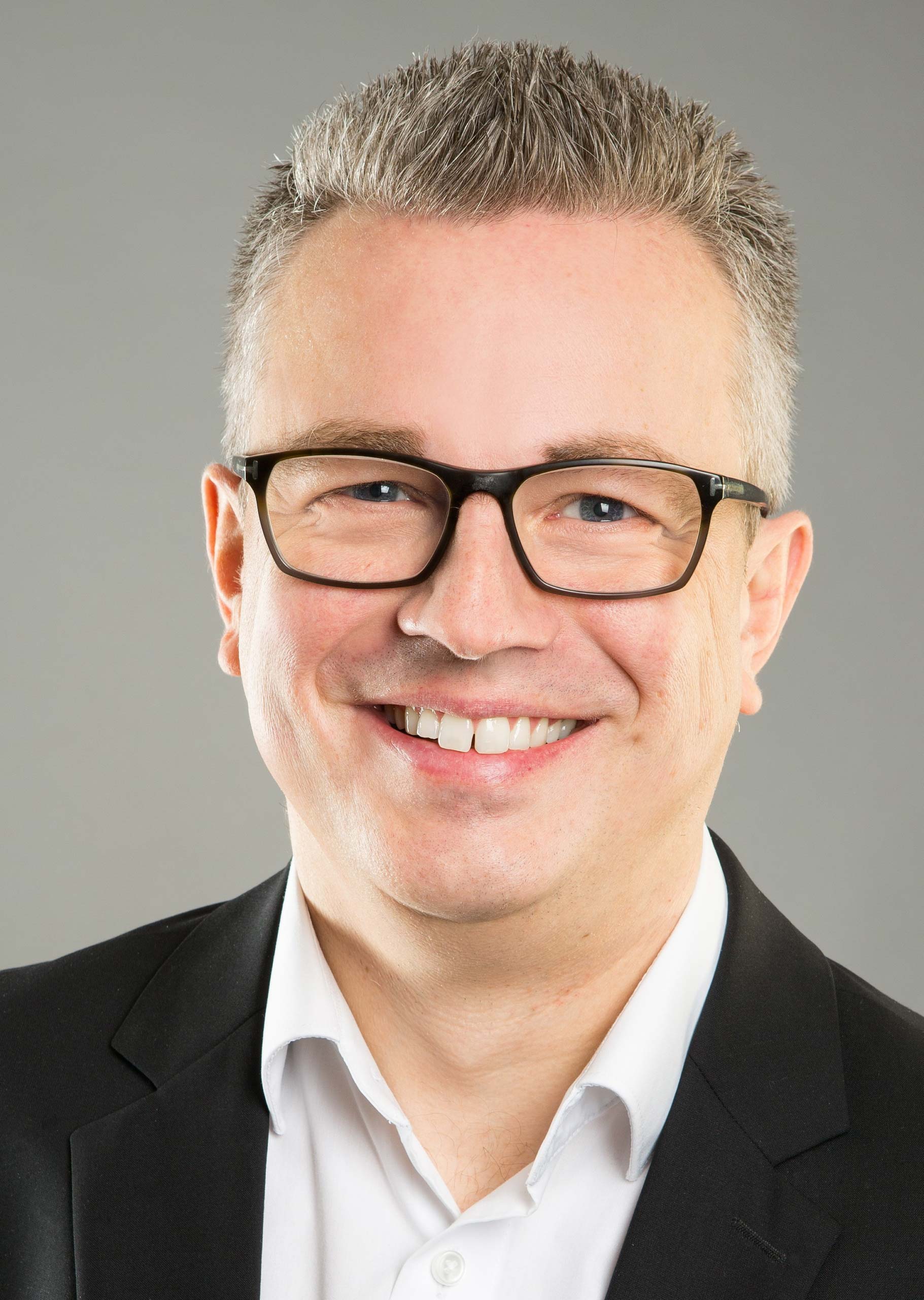 Porträt von Patrick Lamers, lächelnder Mann mit Brille und schwarzem Anzug vor grauem Hintergrund.